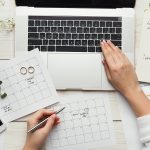 A wedding planner working on their laptop with wedding notes and schedules scattered on the desk with baby's-breath flowers and wedding rings.