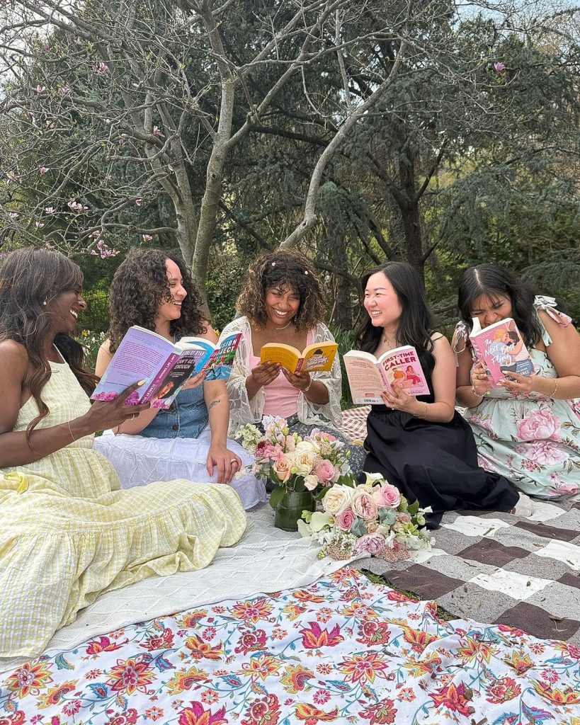 A group of friends sit outside on a picnic blanket reading their book club picks, surrounded by bouquets of flowers and trees.