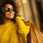 Lady wearing a yellow jumper and sunglasses holding brown paper shopping bags