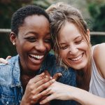 Two women hugging eachother in a close up shot. On the left a black woman wearing a denim jacket and on the right, a caucasion woman wearing a white tank top.