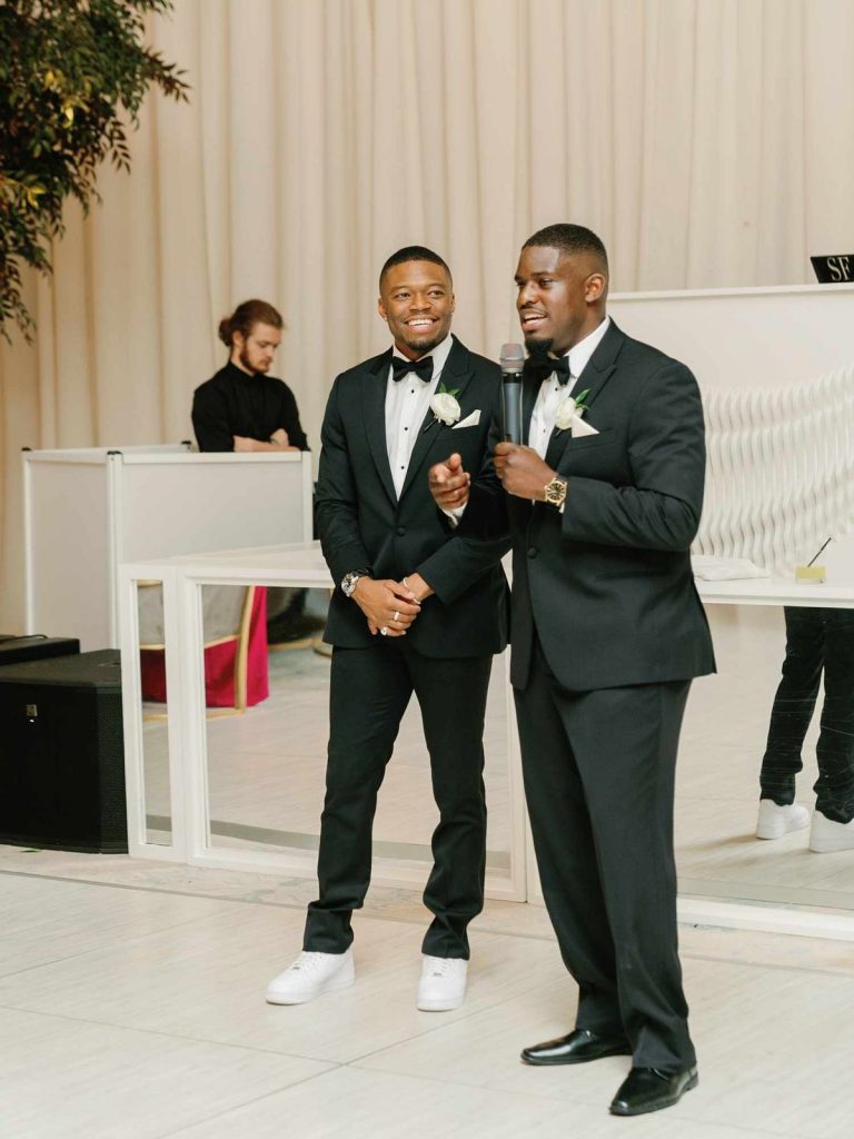A best man giving a toast at a wedding while the groom looks on.