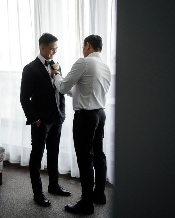 A man is helping the smiling groom get ready by fixing his boutonnière. They are standing next to a large curtained window.