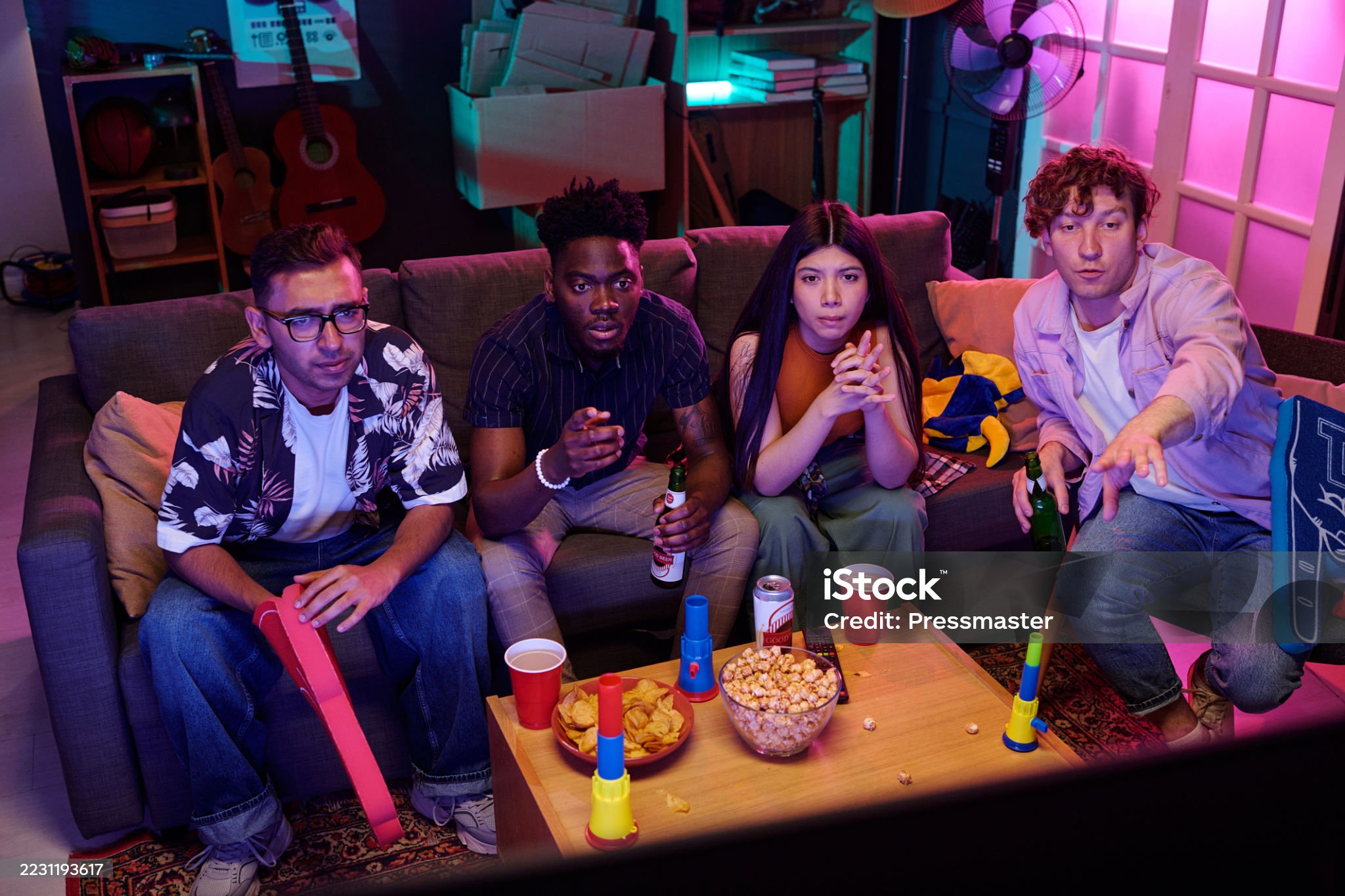 Group of young adults including Caucasian man, Black man, Asian woman, and another Caucasian man watching television together, holding snacks and drinks, sitting on couch in living room