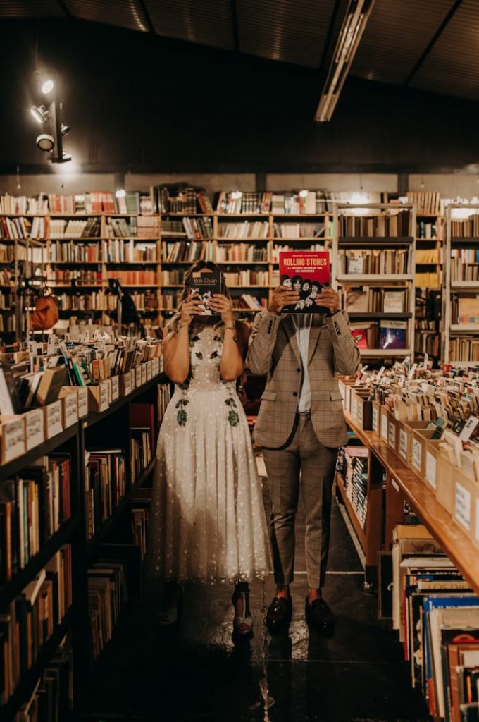 A couple standing in the aisle of a bookshop, holding a book in front of their faces, with bookshelves covering the walls and boxes of books lining the aisle. 