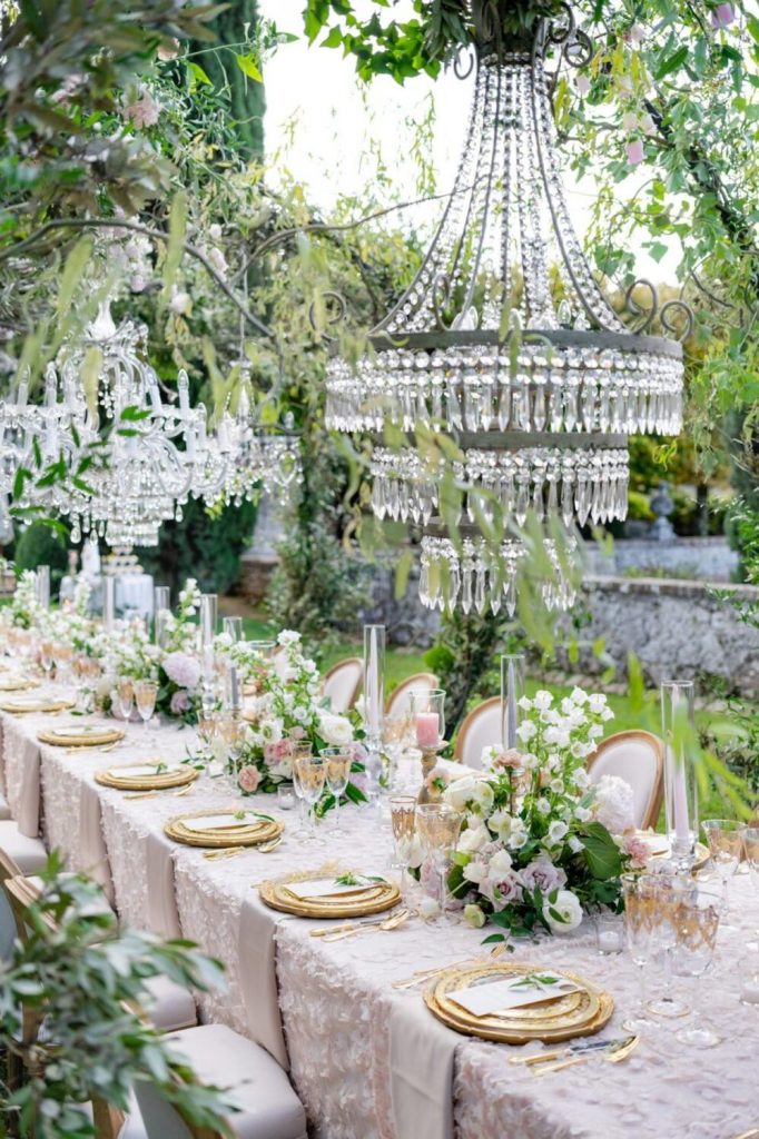 An outdoor wedding reception with decadent decorations, including crystal chandeliers, white and pink roses, and flower appliqué tablecloths.