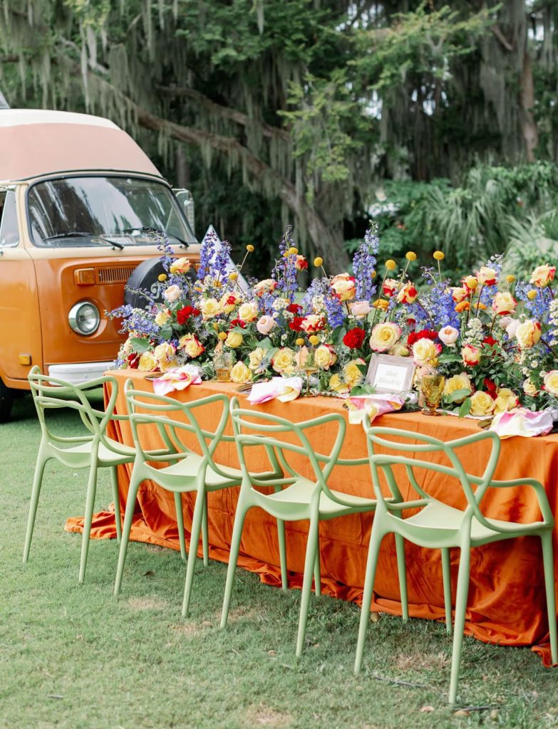 An outdoor table setting with a long, orange tablecloth, a dense flower centrepiece filled with roses, hyacinths, and yellow spring flowers. There are evergreen, mossy trees and a Volkswagen-inspired camper van in the background.