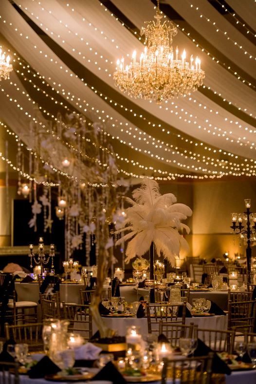 An extravagant ballroom wedding setup, filled with gold chandeliers, candelabras, ostrich feathers, gold and black-themed round tables, and fairy lights.