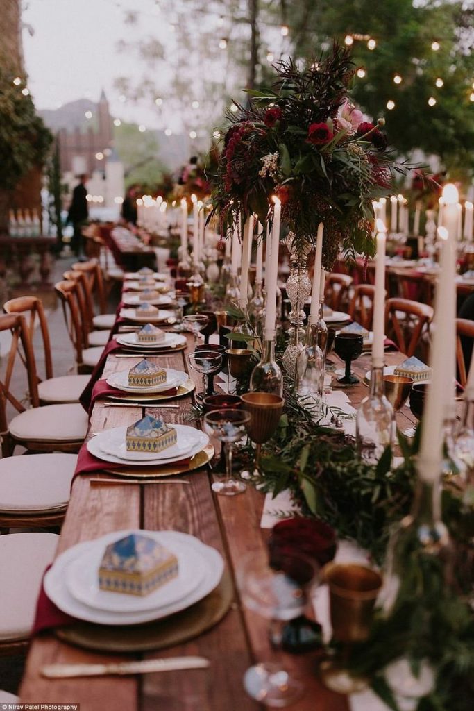 A long rustic wooden table with upcycled glass bottled candle holders placed along a white runner with greenery and rose flower bouquets. String lights are hung above the table, and every plate has a Chocolate Frog box inspired by the Harry Potter book series.