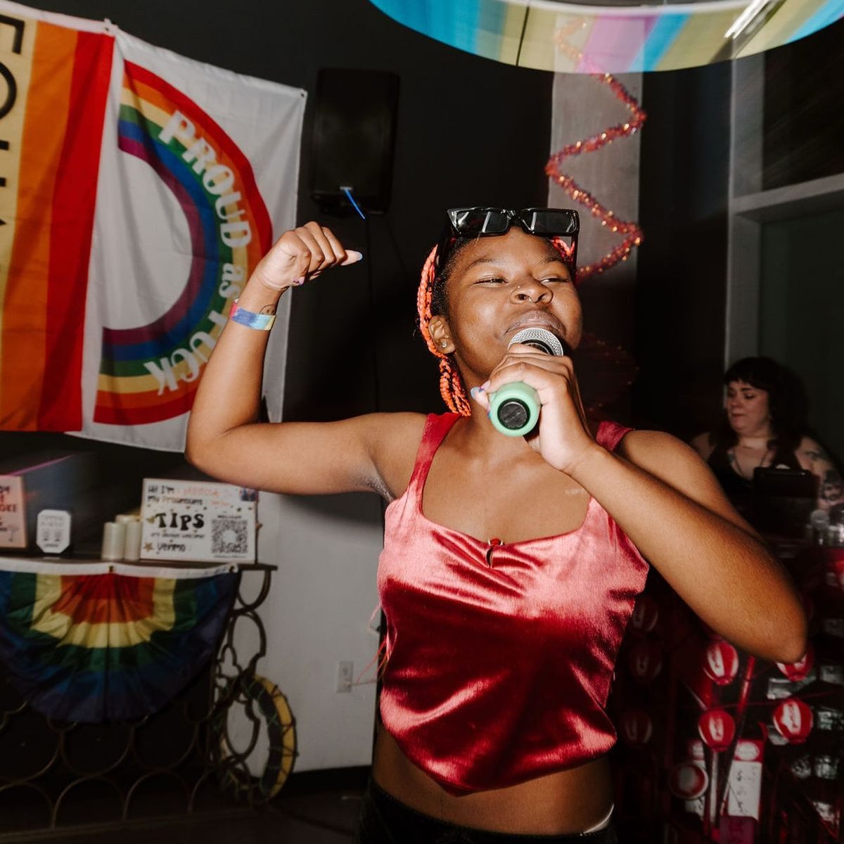 A person singing karaoke wearing sunglasses. In the background there are different styles of pride flags and decorations on display.