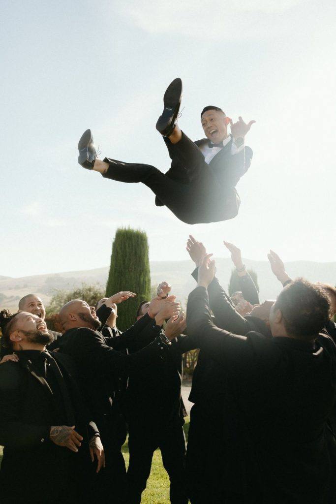 A picture of a happy groom being thrown up into the air and caught by multiple groomsmen.