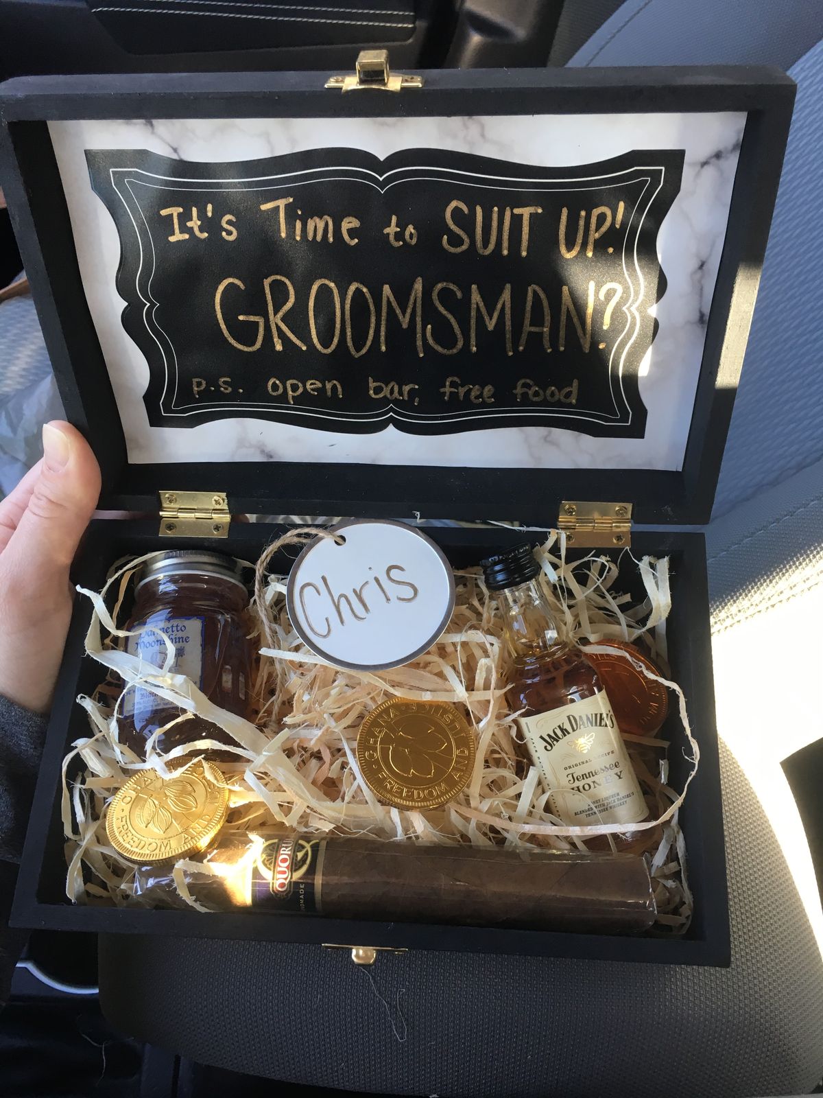 A groomsman gift box with a proposal message, a name tag, and a selection of gifts, including a cigar and bottle of whiskey.