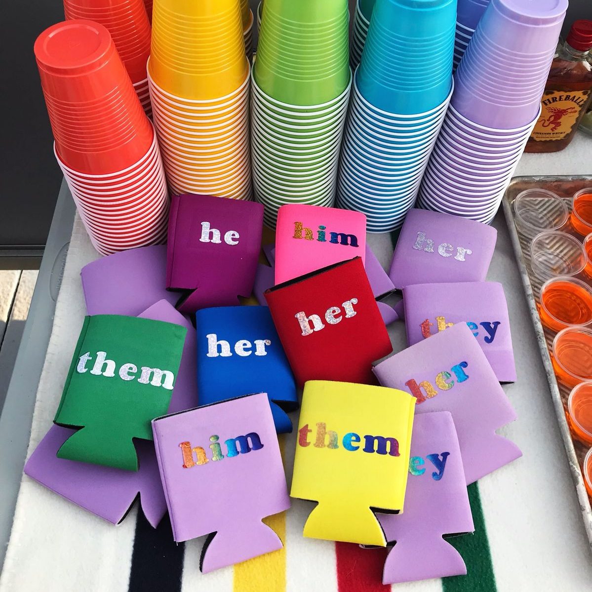 Rainbow-colored plastic party cups and colorful can coolers with pronoun text written on them set down on a table. 