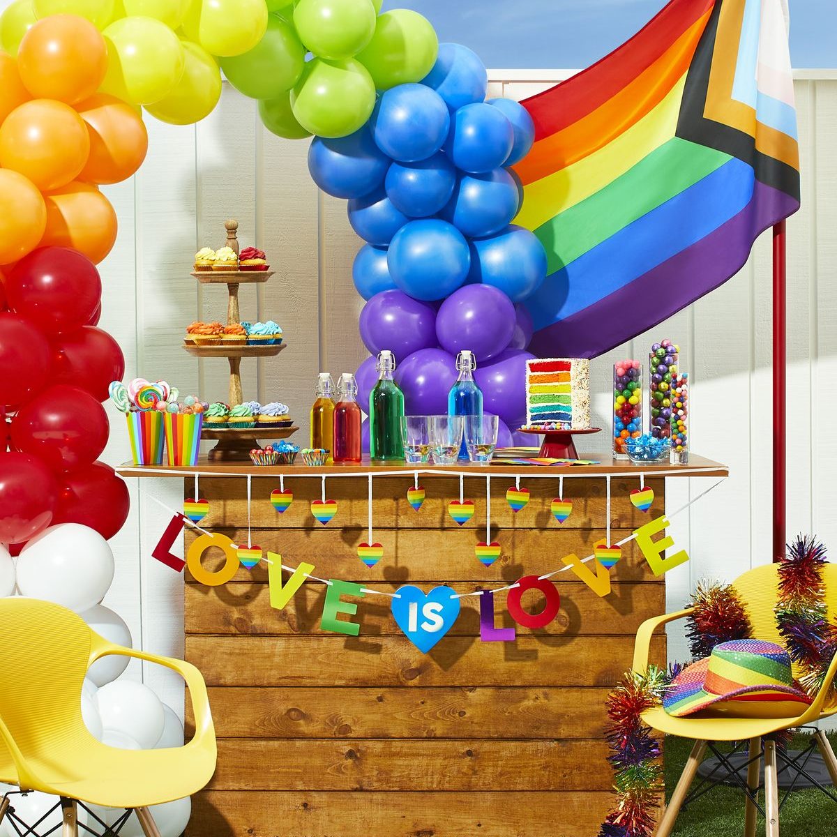 A backyard pride party table set up with colorful decorations, drinks and candy. In the background there is a rainbow balloon arch and a pride flag as well as two bright yellow chairs.