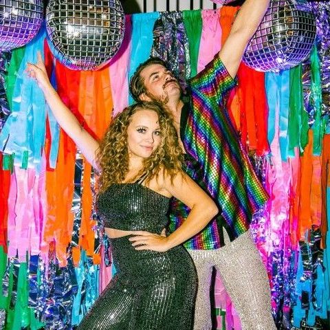 Two friends posing with their arms up in front of a colorful tissue paper-style backdrop and silver disco balls.