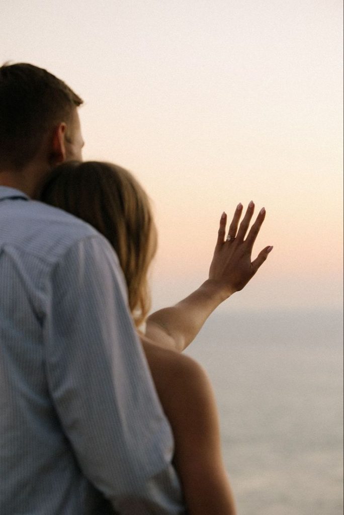 An engaged couple standing together at sunset after the big question was answered! They look out at the sunset and admire the ring that one of them is holding up to the sky.