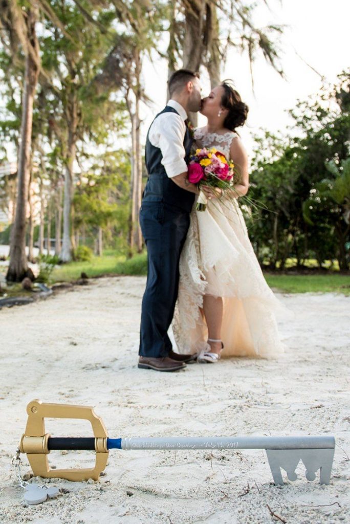 A bride and groom share a kiss outdoors with a Kingdom Hearts prop lying on the ground in front of them.