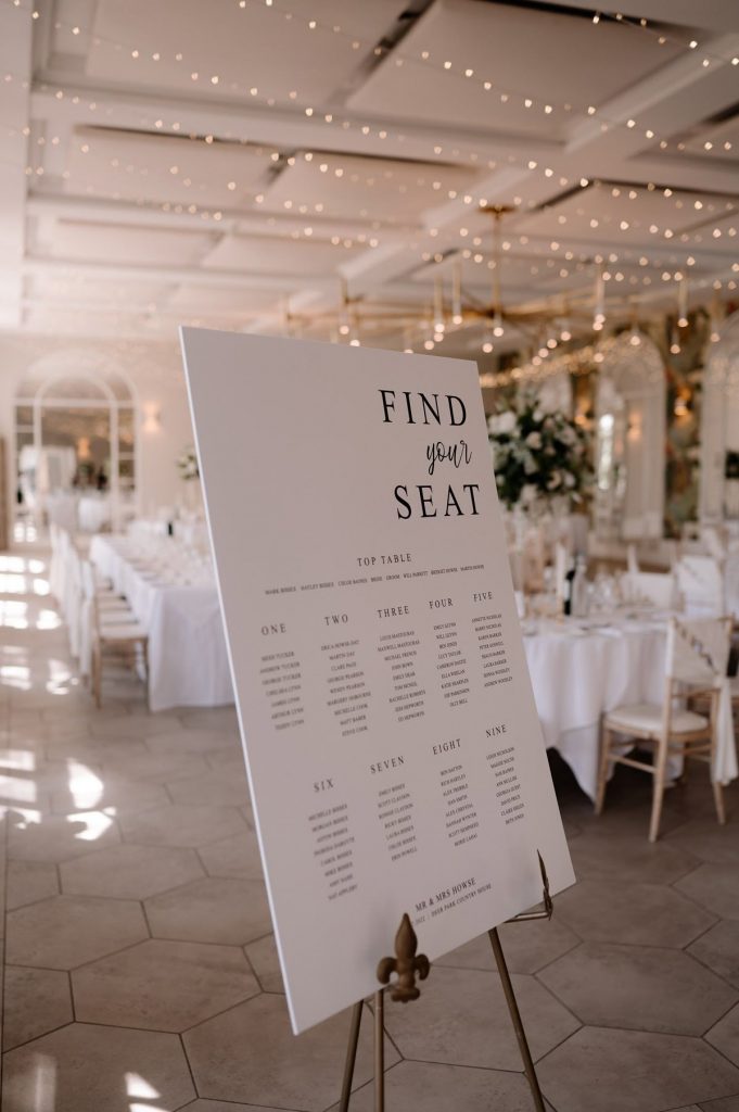 A beautiful wedding seating chart situated in the dining room, which is decorated with fairy lights and white decorations.