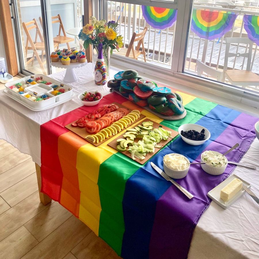 A large table with a rainbow table cloth with lots of colorful fruit and vegetables, bagels, cupcakes, as well as various spreads.