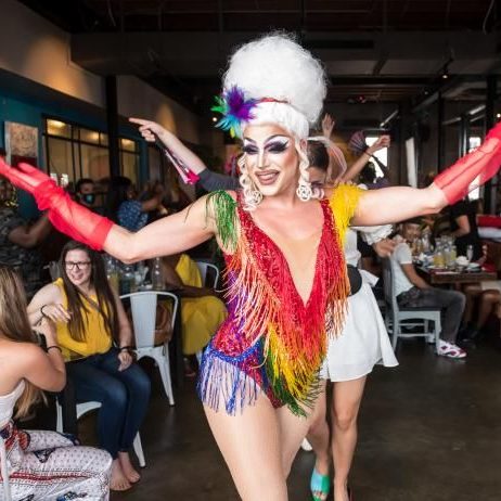 A drag queen dressed in a rainbow bodysuit performing for a crowd in a brunch setting. 
