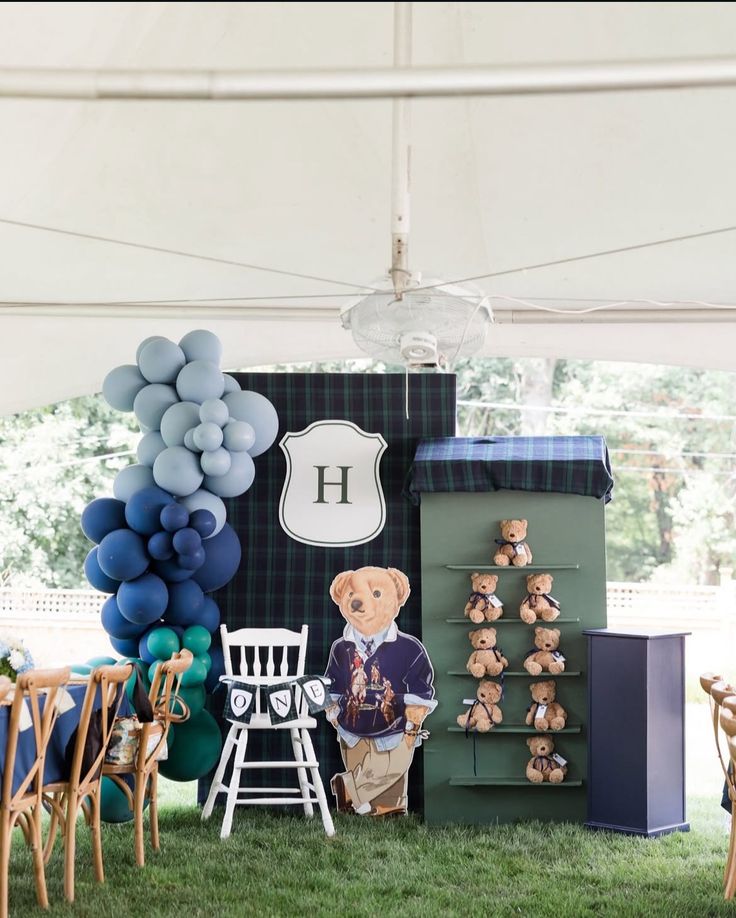 Outdoor setup with a blue balloon arch. A plaid backdrop and monogrammed sign and a teddy bear arch in soft, neutral colors.