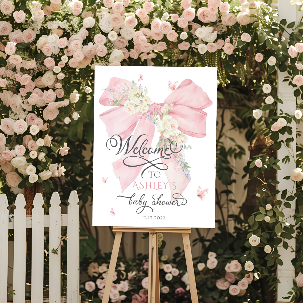 A pink bow baby shower welcome sign with watercolor floral design and elegant calligraphy on white background, displayed on wooden easel against lush pink and white rose flower wall with white picket fence.