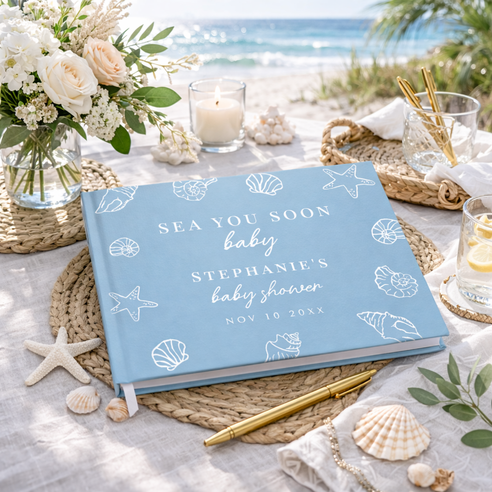 A Sea You Soon nautical baby shower guest book in dusty blue with white seashell and starfish illustrations, displayed on beach-themed tablescape with ocean view, flowers, candles, and coastal decor.