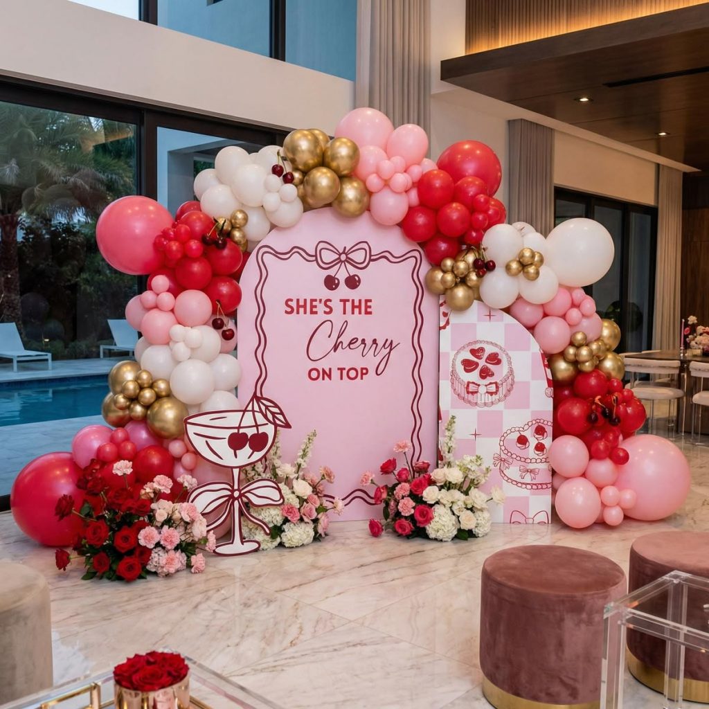A mix of red, white, pink, and gold balloons are hung around a cherry-themed sign. Furniture can be seen in the foreground and a swimming pool can be seen in the background.