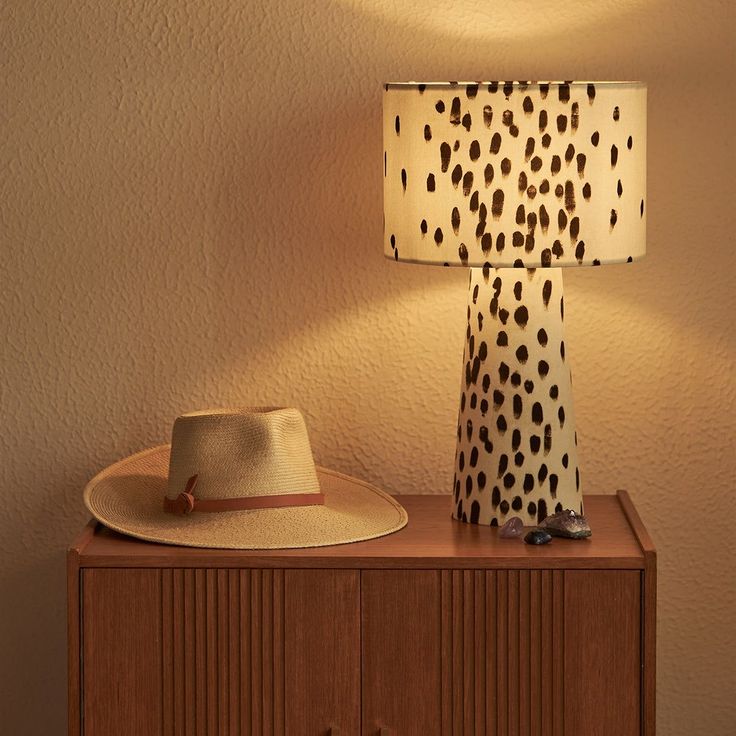 A black and white spotted table lamp is shown on a brown side table. There is also a cream and brown hat to the left on the table.