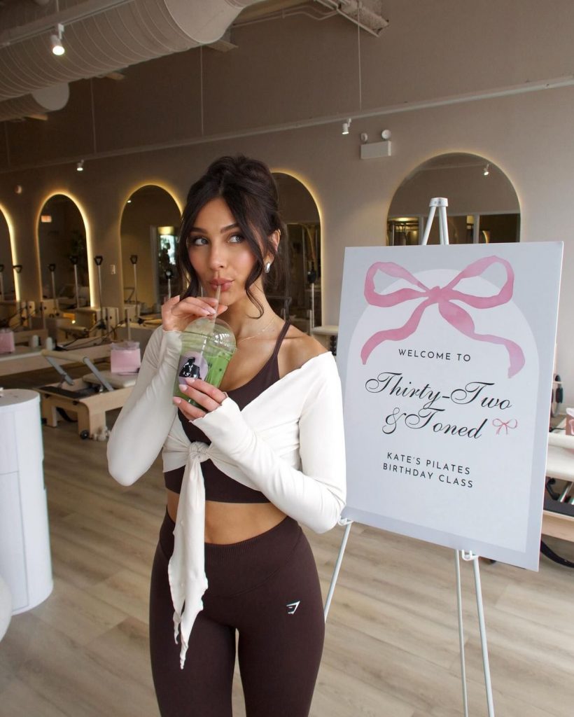 A woman in gym gear is standing next to a sign announcing an exercise class for her birthday. Gym equipment and large mirrors can be seen in the background, and the woman is drinking matcha tea through a straw.