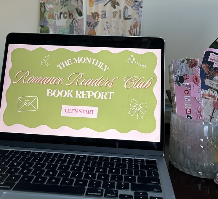 A laptop shows off a monthly book report for a book club. The laptop is sitting on a table surrounded by bookish crafts.