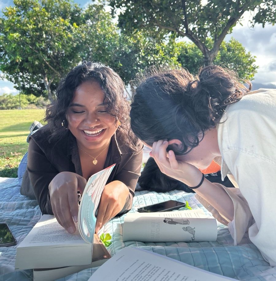 Two friends are lying down on a blanket outside, laughing about the books that they are reading together.