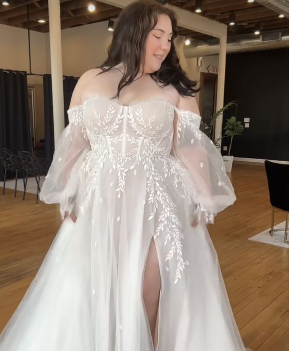 a beautiful woman displaying an off the shoulder, long sleeve, white sheer wedding dress with a slit up the right thigh. She is standing in a living room-like scene with a wooden floor, black walls and furniture in the background.