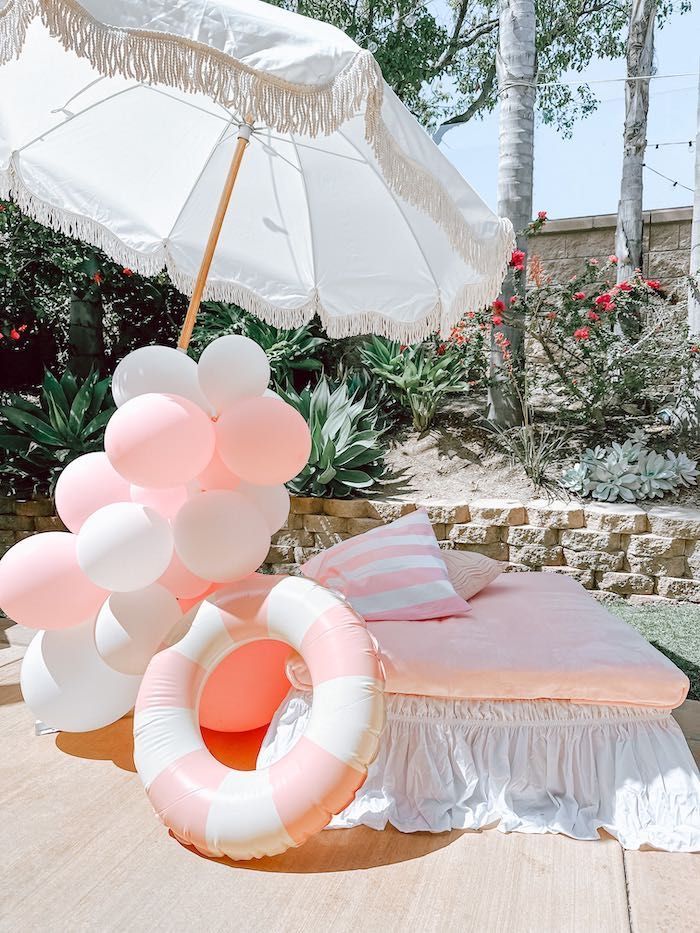 A parasol, life buoy, and pink and white balloons are shown in an outdoor setting. A striped pillow and blanket are lying on top of a sun lounger.