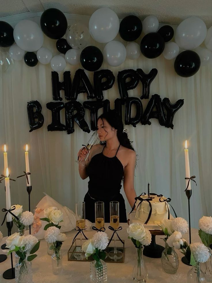 A woman wearing a black dress is taking a drink from a champagne flute. Black and white balloons are hanging above her, and a birthday banner is hanging behind her.