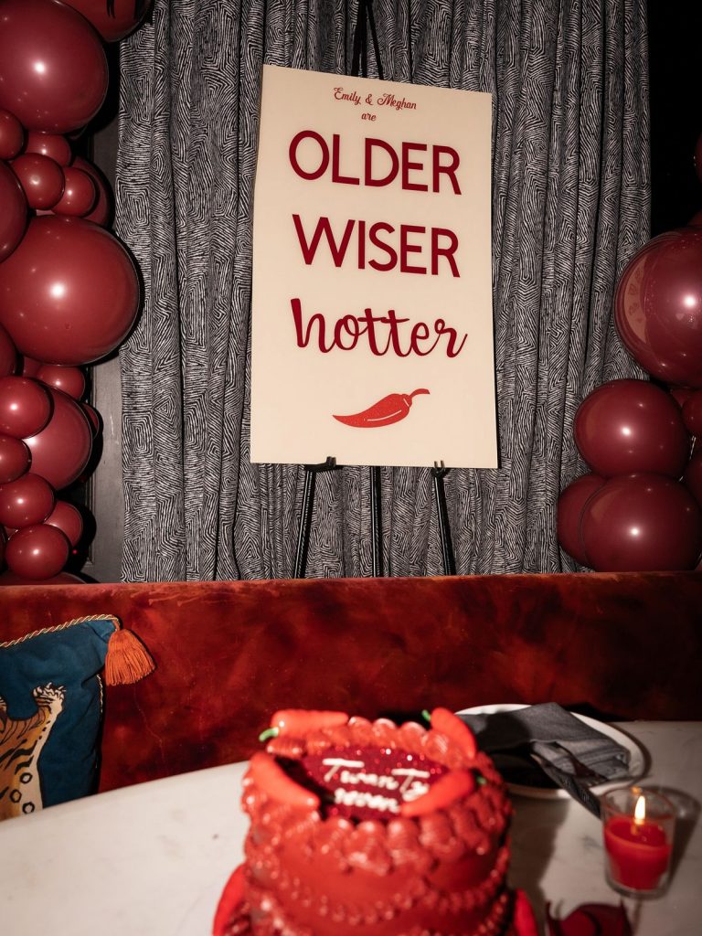 A red cake is on a table in the foreground. In the background there is a sign with a spicy pepper theme, red balloons, and a gray curtain.