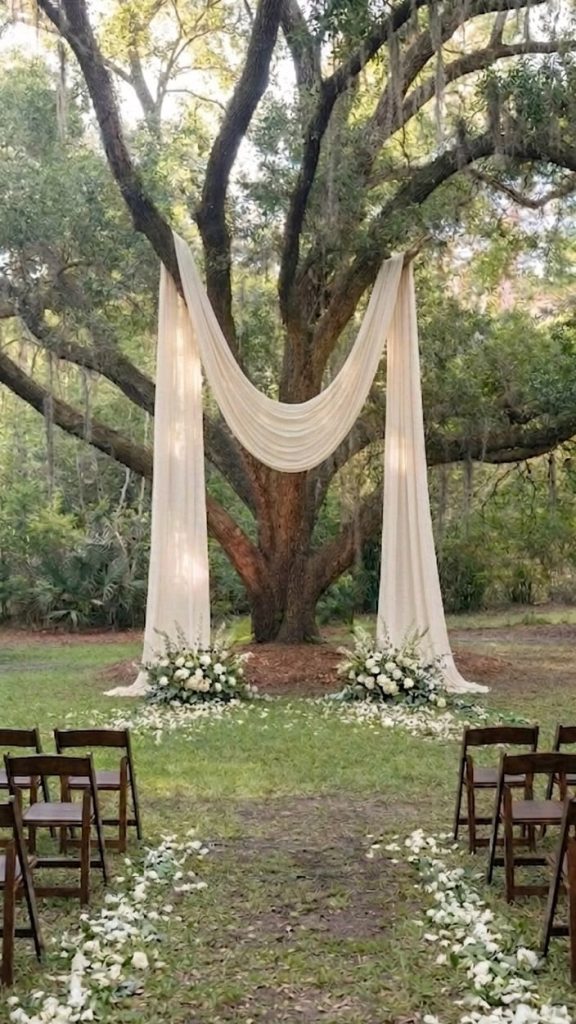 A dark wooden tree with several large branches with an off-white lined draped over forming an arch. At either side of the arch there is a white bouquet of flowers and white petals surrounded by a green area.