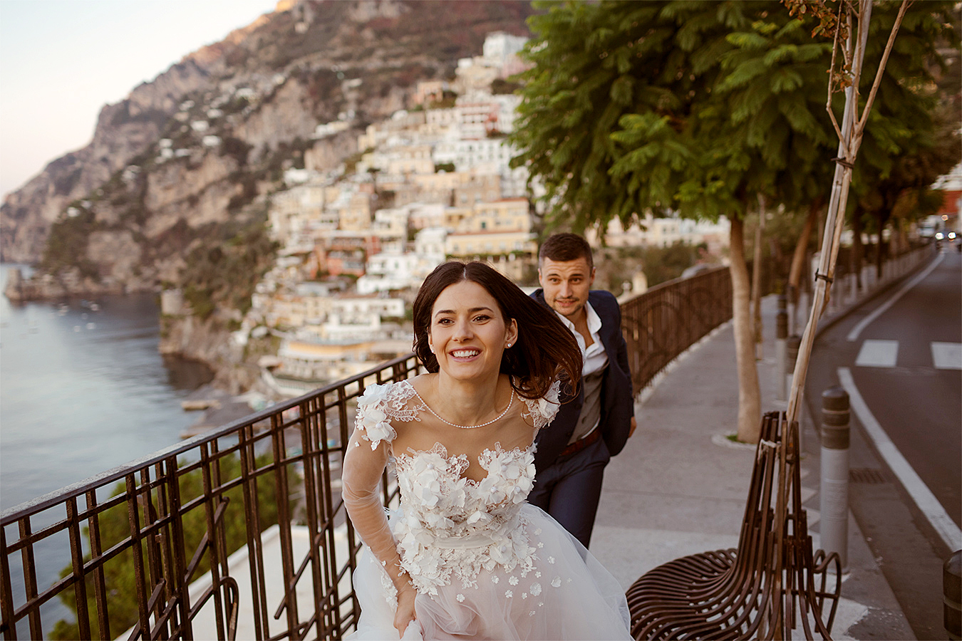 A smiling bride and groom are running down a street in a European location.
