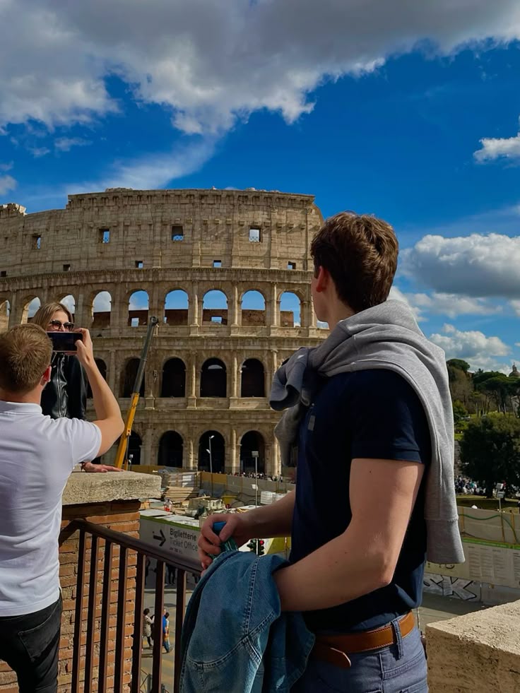 A man with his back turned wearing a t-shirt and jeans looking at the Colosseum in Rome. In the background there are two people taking photos.