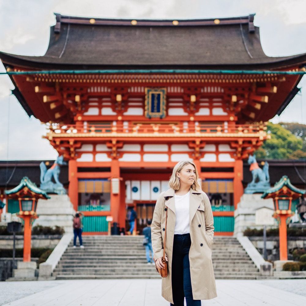 A woman wearing a cream coat and navy pants stands in front of a large traditional Japanese shrine.