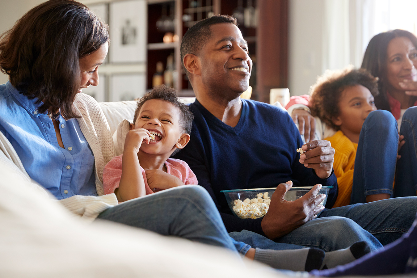 A family watch movies on the sofa while eating popcorn and laughing.
