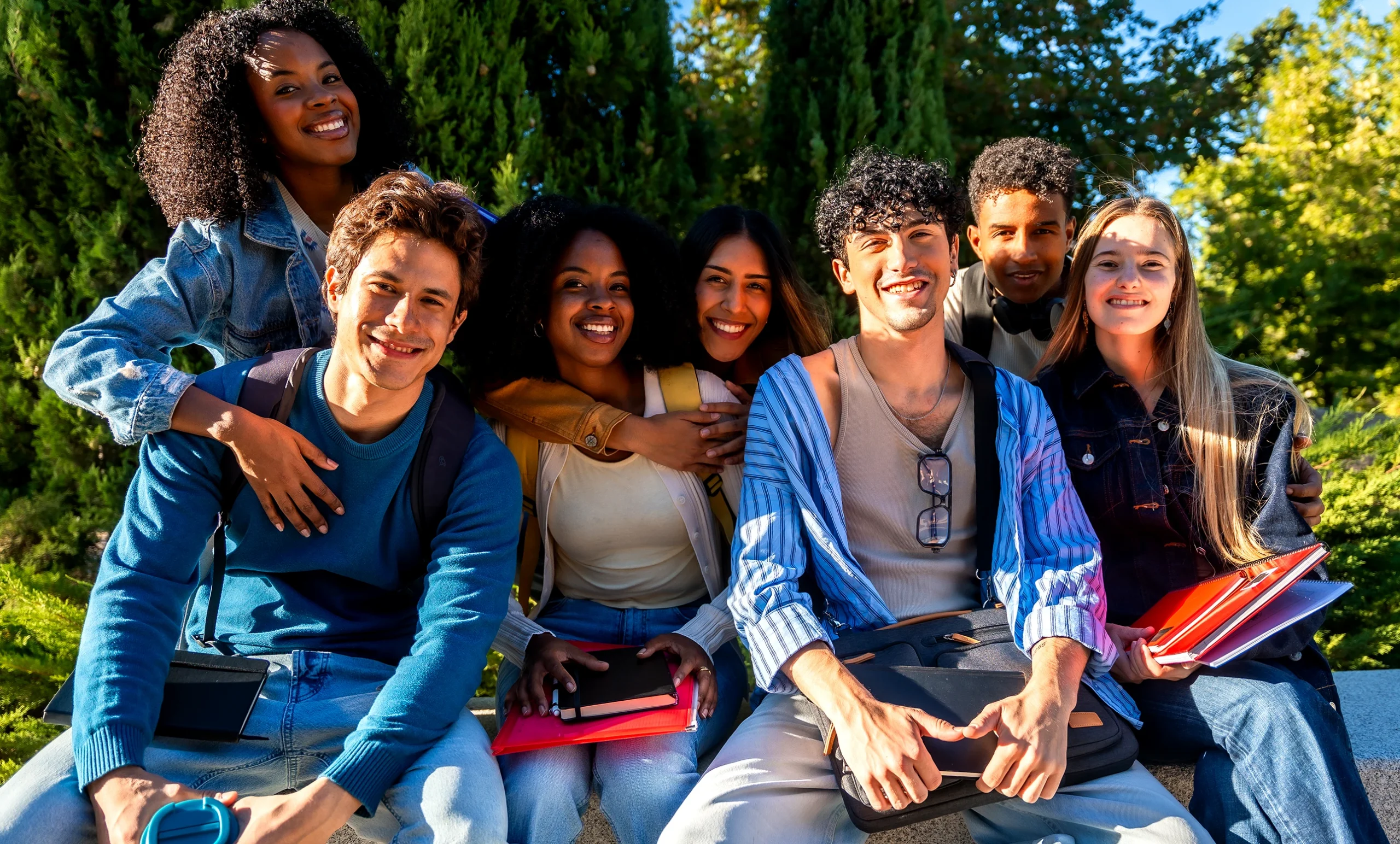 A group of seven diverse university students sit closely together on an outdoor stone bench on campus. They are smiling broadly at the camera under bright sunlight. Most of the students hold colorful planners and textbooks, and several wear backpacks. All getting ready for their first day back to school!