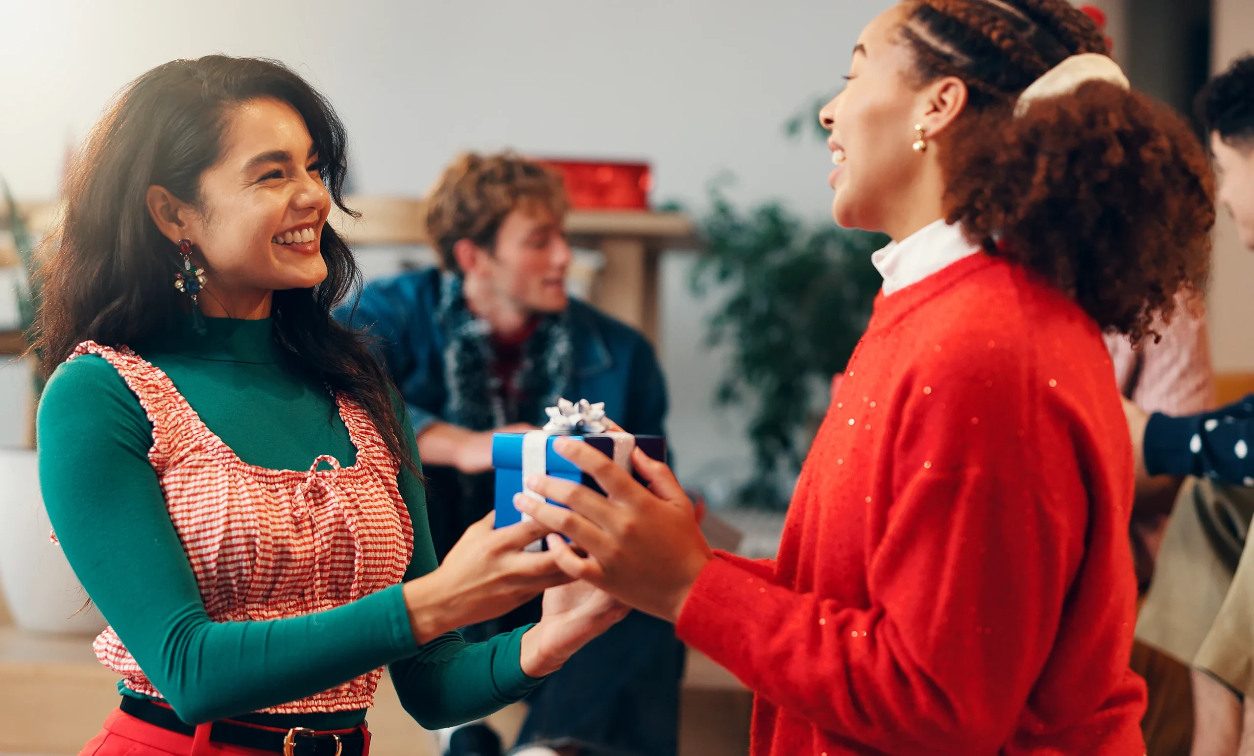 A smiling woman in a green top and a red-and-white gingham dress hands a small blue gift box to another woman wearing a red sweater at a festive gathering, while other people mingle in the background.